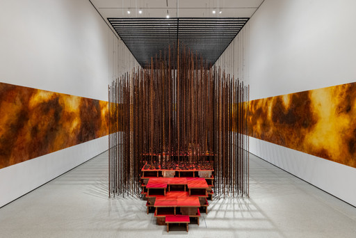 The image shows a room with predominantly white and fiery orange-yellow walls. Centered is a structure made of vertical rods hanging down, encompassing stacked red platforms. This is an AI generated description. To give feedback, email ai@moma.org.